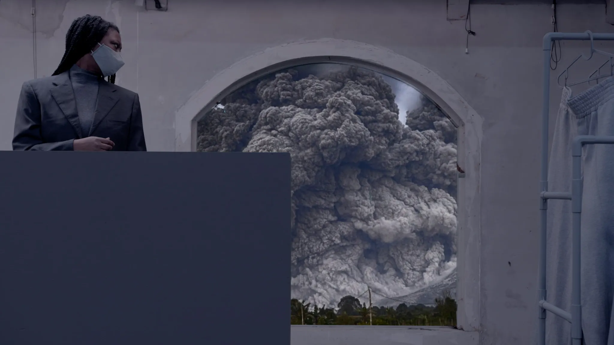 A character with black braids, attired in a sophisticated black suit and wearing a face mask, glances over their shoulder toward a window framing a landscape of catastrophic turmoil. The landscape features thick, volcano-like black smoke, setting a dramatic tone.