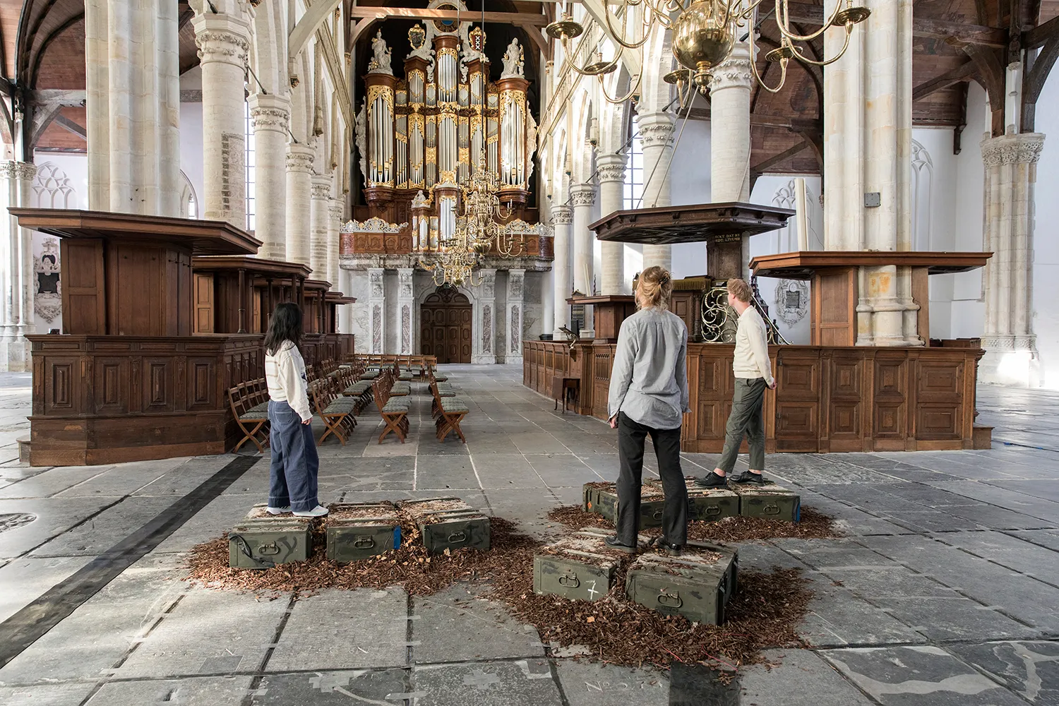 Three individuals standing on green wooden boxes placed on wood chips, while staring towards the church organ inside the Oude Kerk