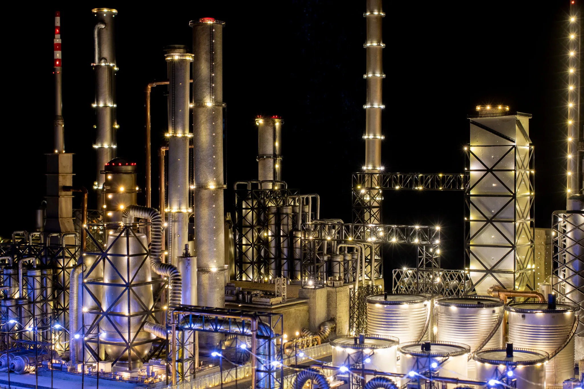 Een olieraffinaderij-industriegebied 's nachts, met helder verlichte witte torens en cilinders in wit en blauw licht.