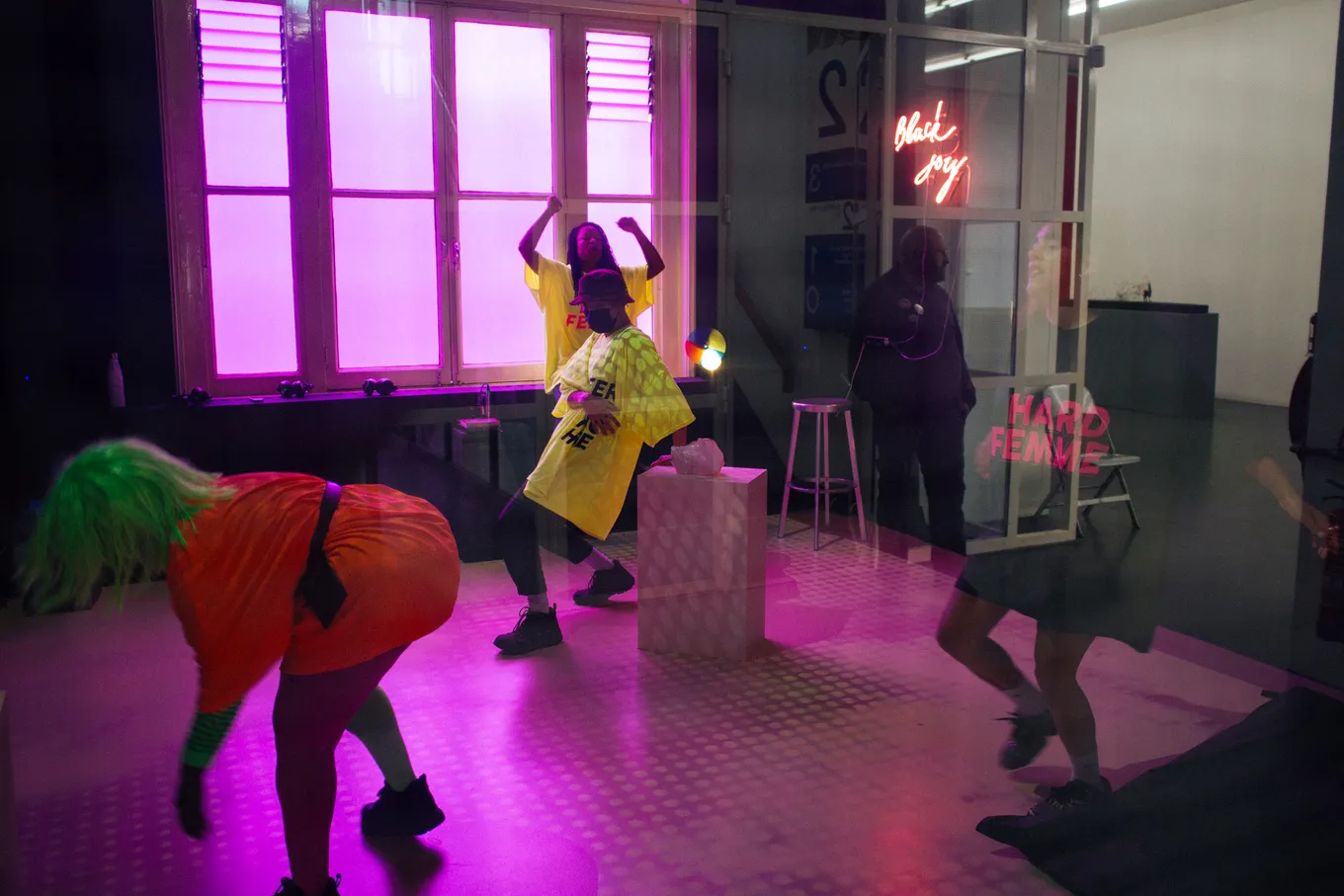 Performers dancing in the exhibition space, with a 'Black Joy' neon light on the back wall. A performer wears a tshirt with the text 'Hard Femme'.