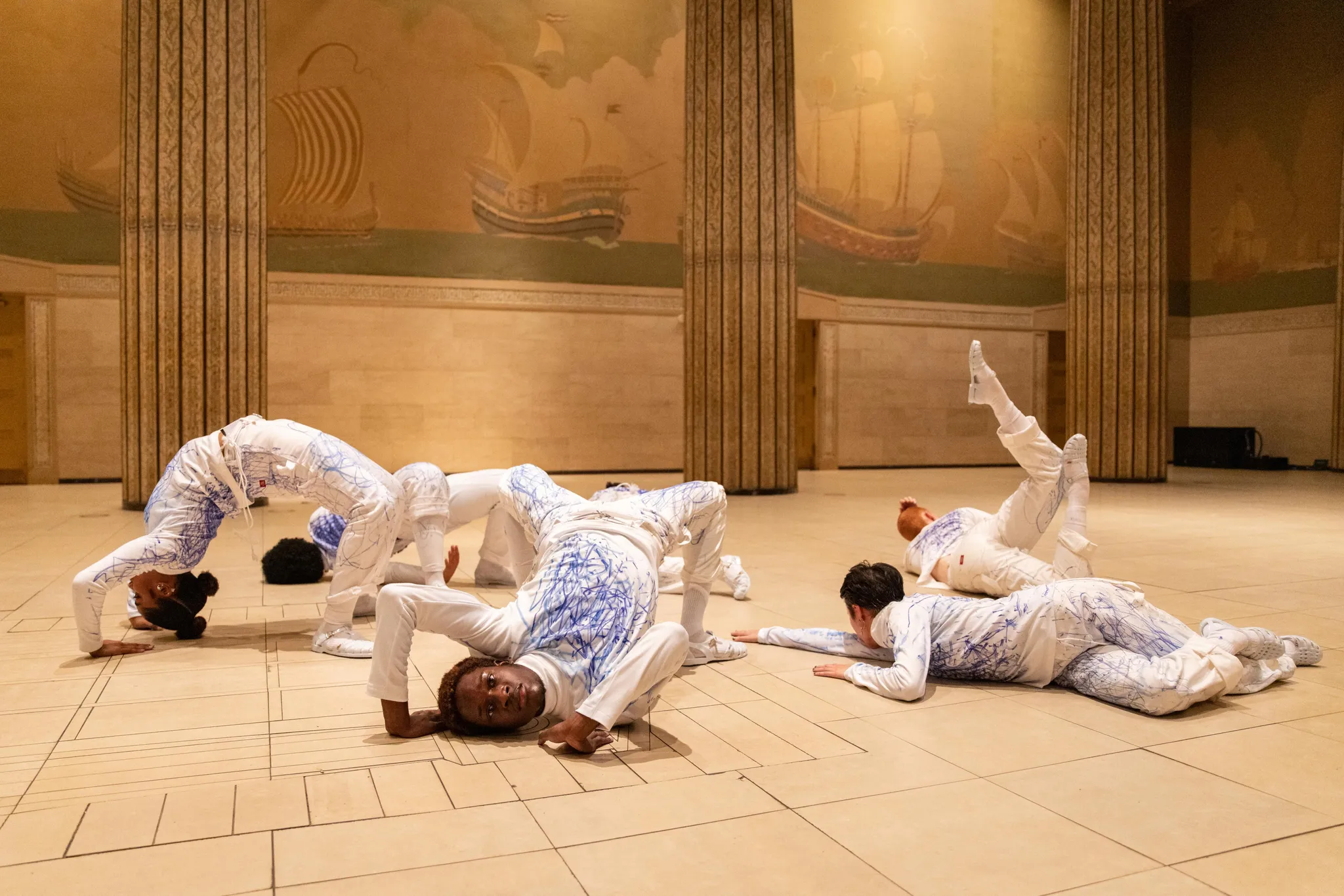 Six performers in assorted poses on the ballroom floor within a spacious environment adorned with pillars and wall paintings portraying ships