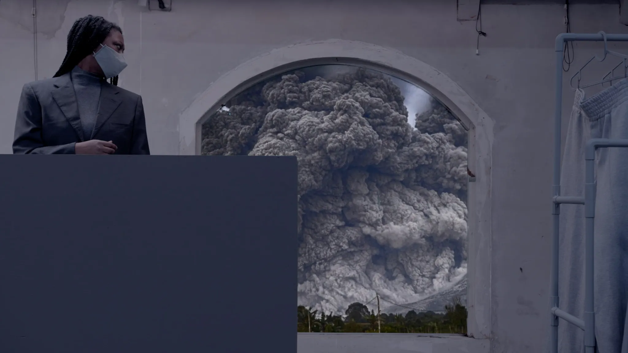 Een personage met zwarte vlechten, gekleed in een verfijnd zwart pak en met een gezichtsmasker, werpt een blik over hun schouder naar een raam dat een landschap van catastrofale chaos omlijst. Het landschap toont dikke, vulkaanachtige zwarte rook, wat een dramatische sfeer creëert.