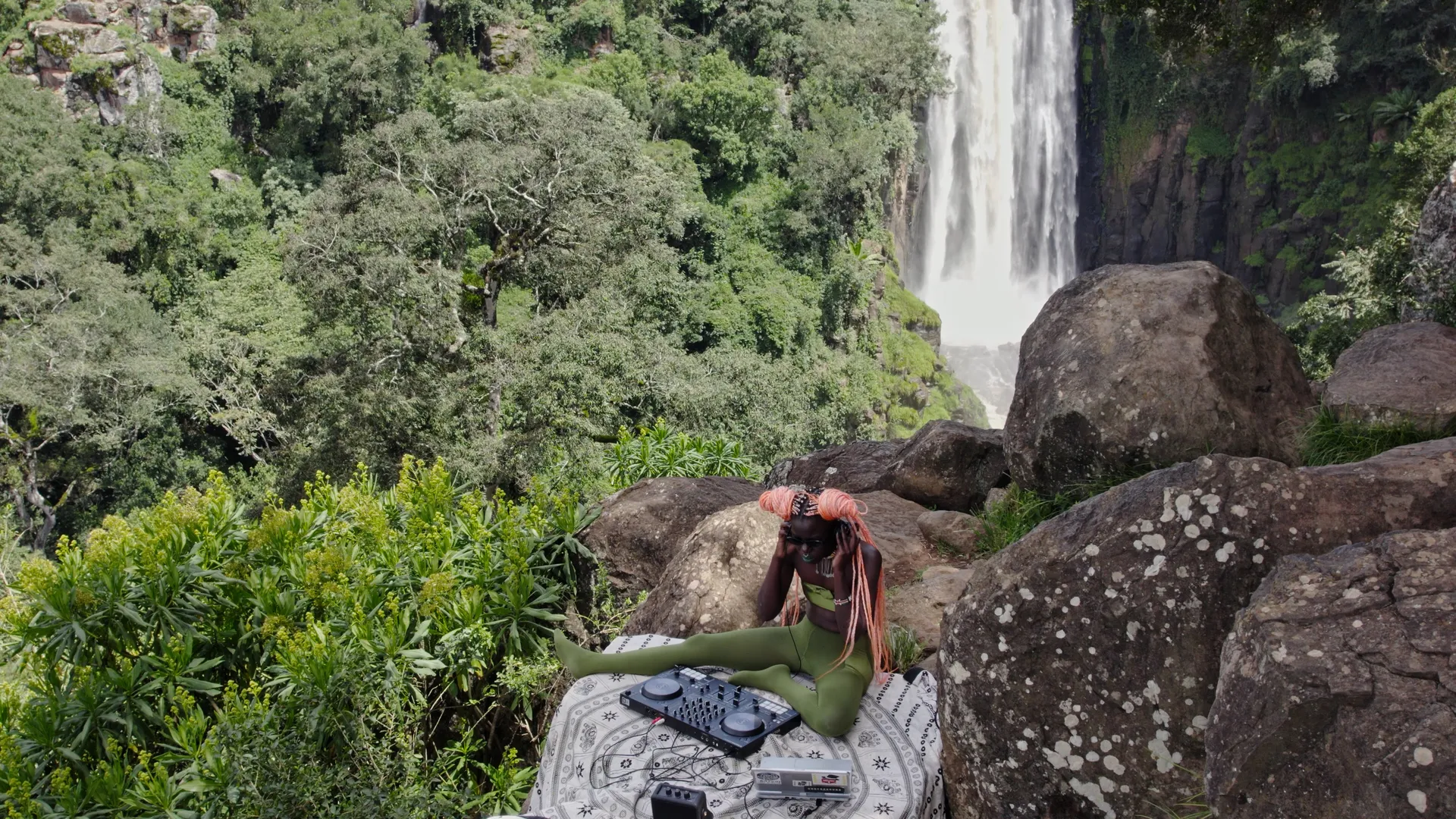 A person with dark skin and pink braided hair listening to a track on their headphones, sitting on a rock with mixing table & laptop in front of a lush background with waterfall