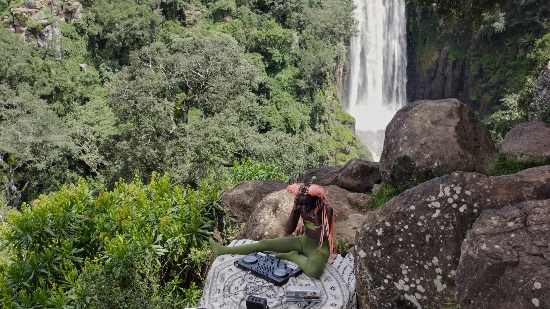 Een persoon met donkere huid en roze gevlochten haar luistert naar een track op koptelefoon, zittend op een rots met mengtafel & laptop voor een weelderige achtergrond met waterval