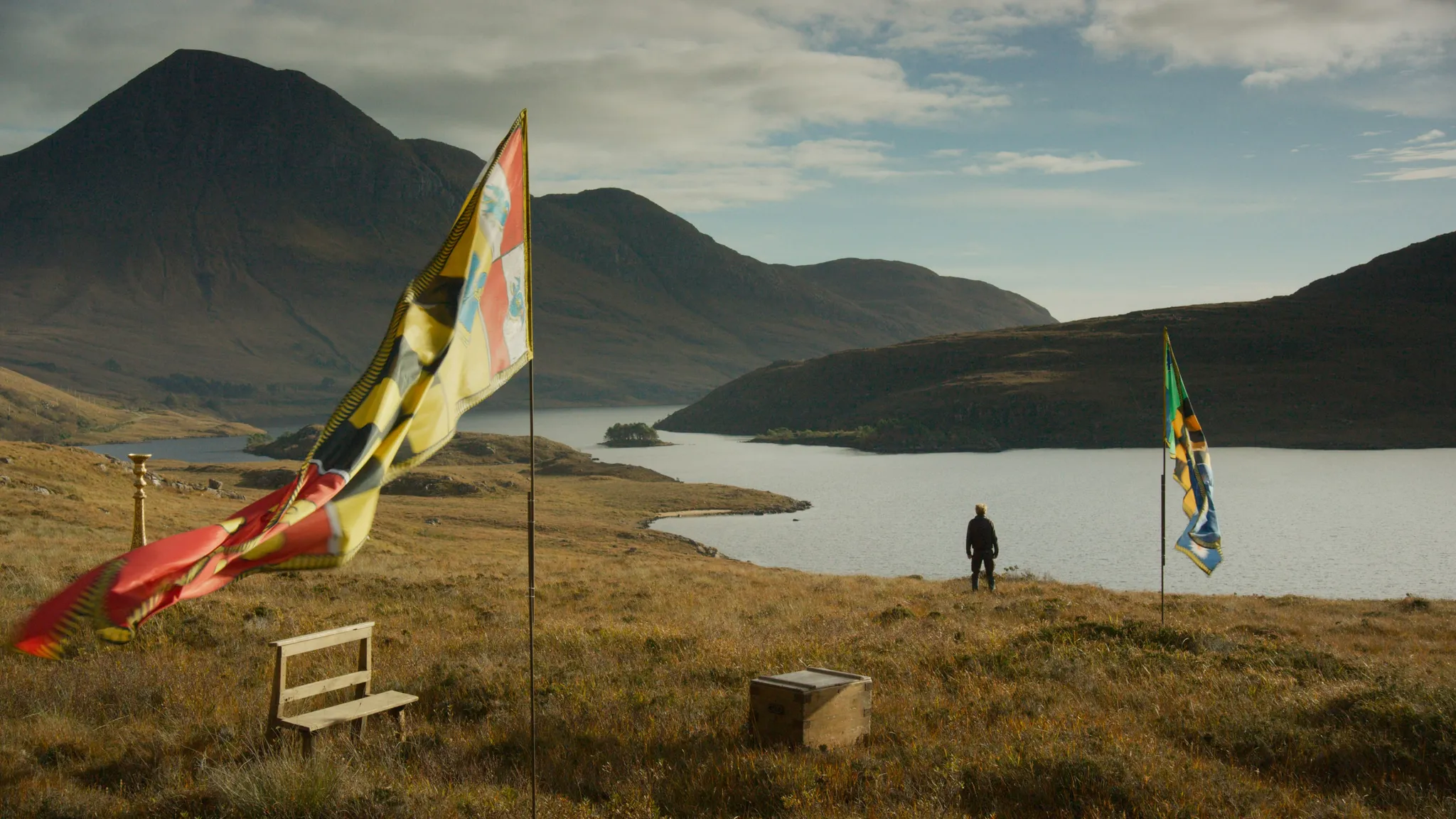 A mountainous landscape with a lake (or river), two undefined flags planted in the ground and a person staring at the water