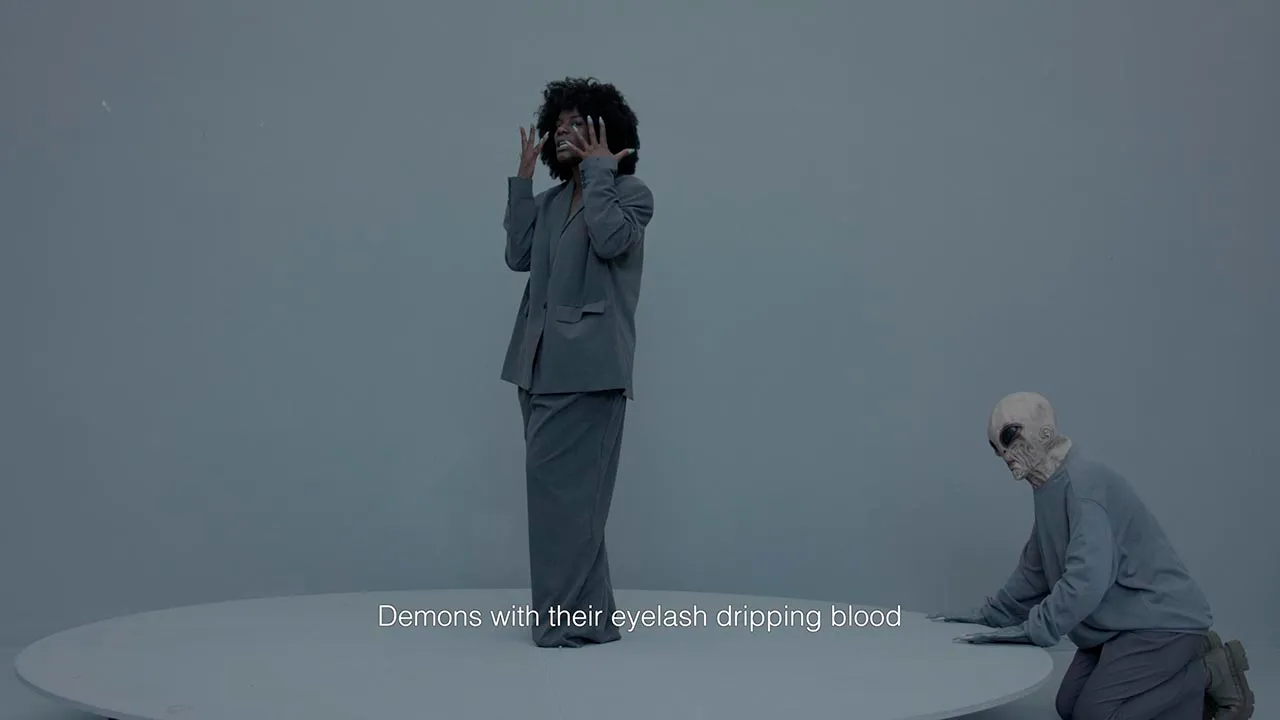In a grey setting, a Black woman, wearing a loose grey tuxedo, raises her hands in front of her face while an alien figure emerges stealthily from behind. The caption reads 'Demons with their eyelashes dripping blood’