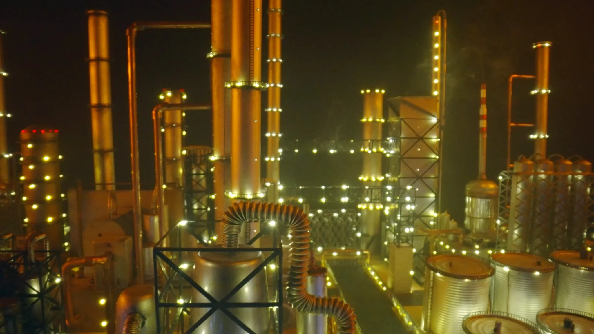 An oil refinery industrial zone aglow at night, bathed in an ambient orange hue amidst a smog-laden atmosphere.