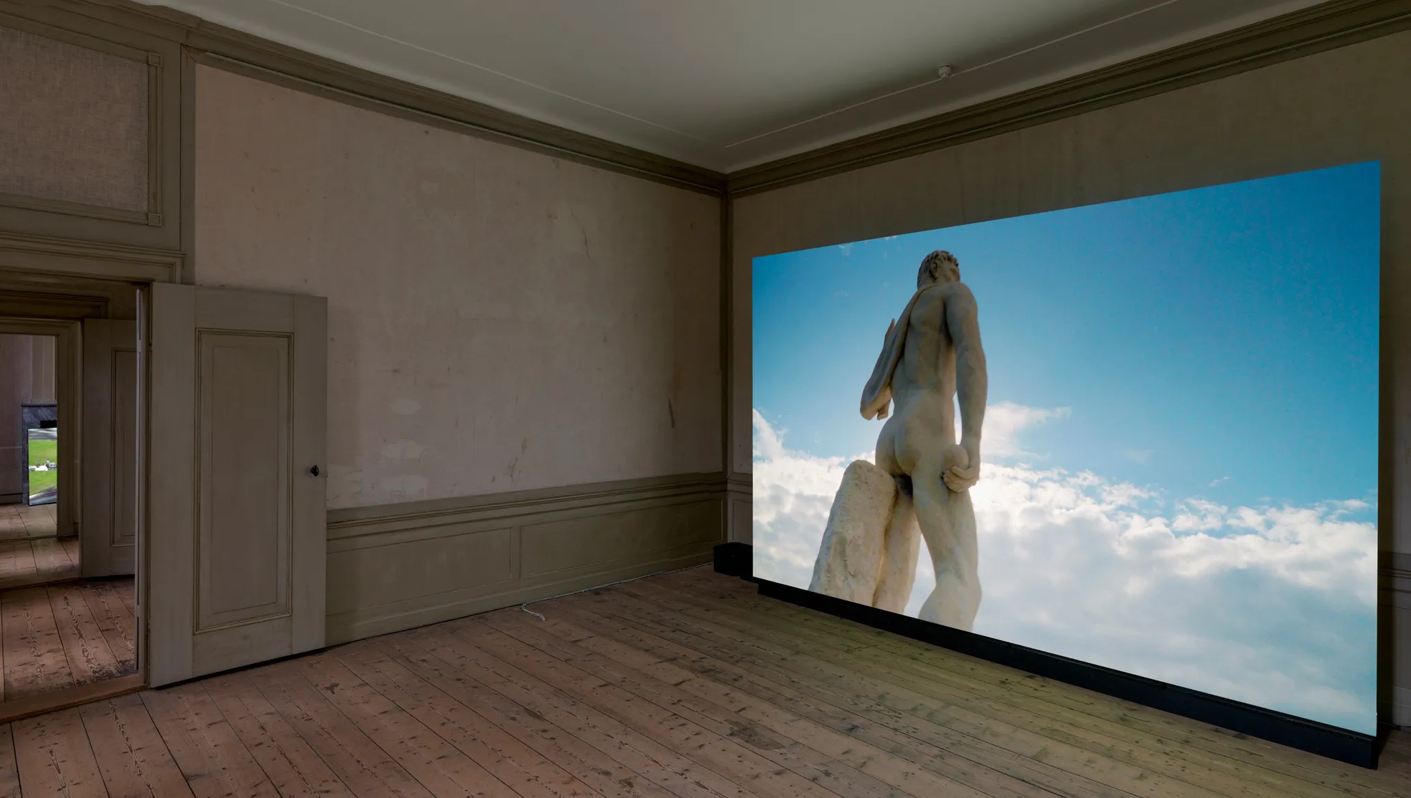 Video on a large screen depicting the backside of a tall classical statue of the Stadio dei Marmi in front of a blue, partly cloudy sky