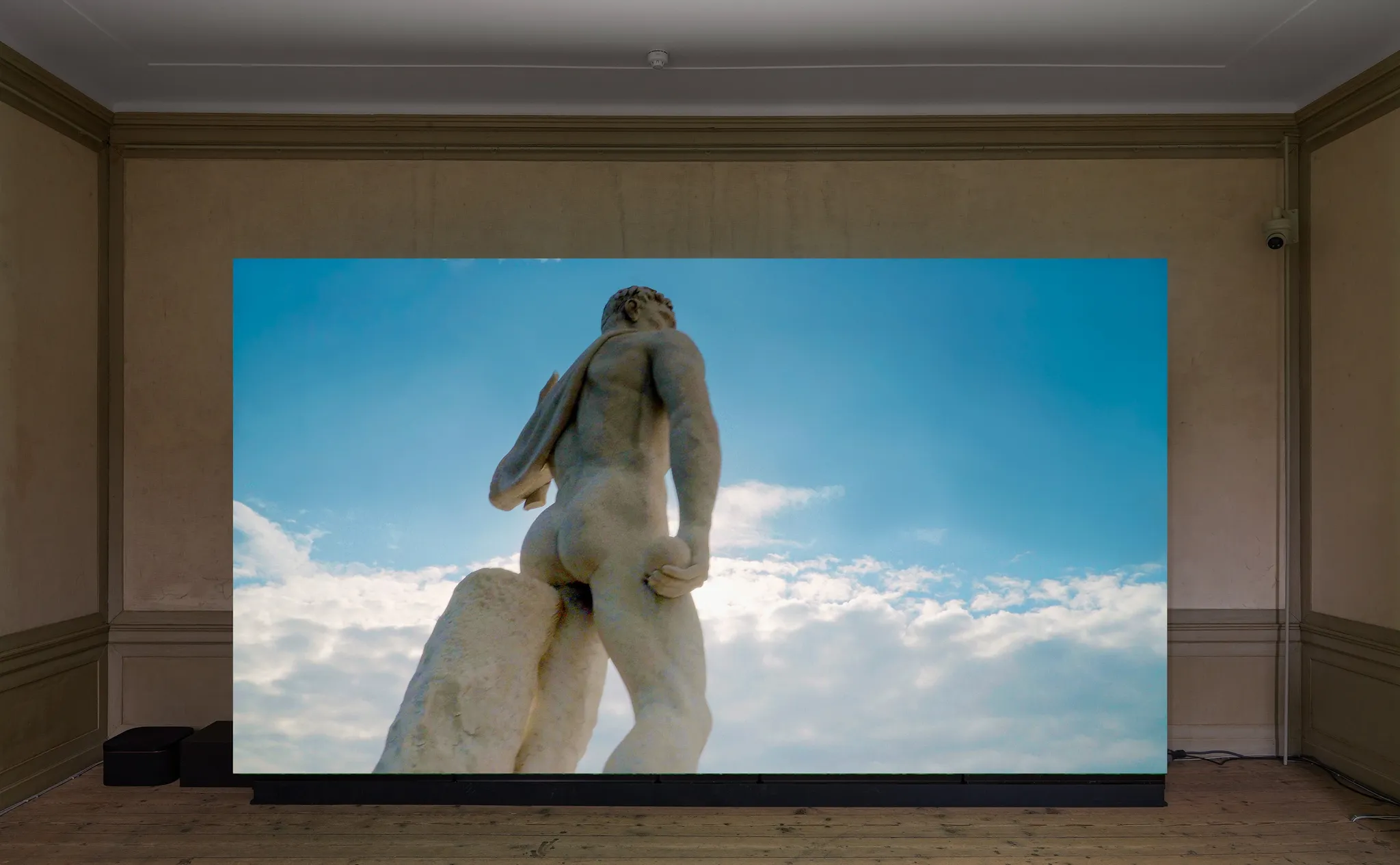 Video on a large screen depicting the backside of a tall classical statue of the Stadio dei Marmi in front of a blue, partly cloudy sky