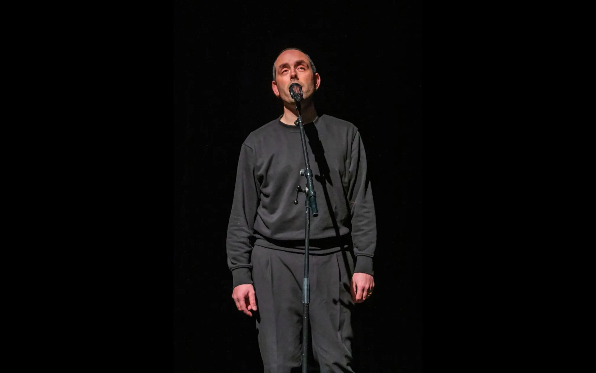 Ed Atkins reciting in front of a microphone, wearing black clothes and with shaved head