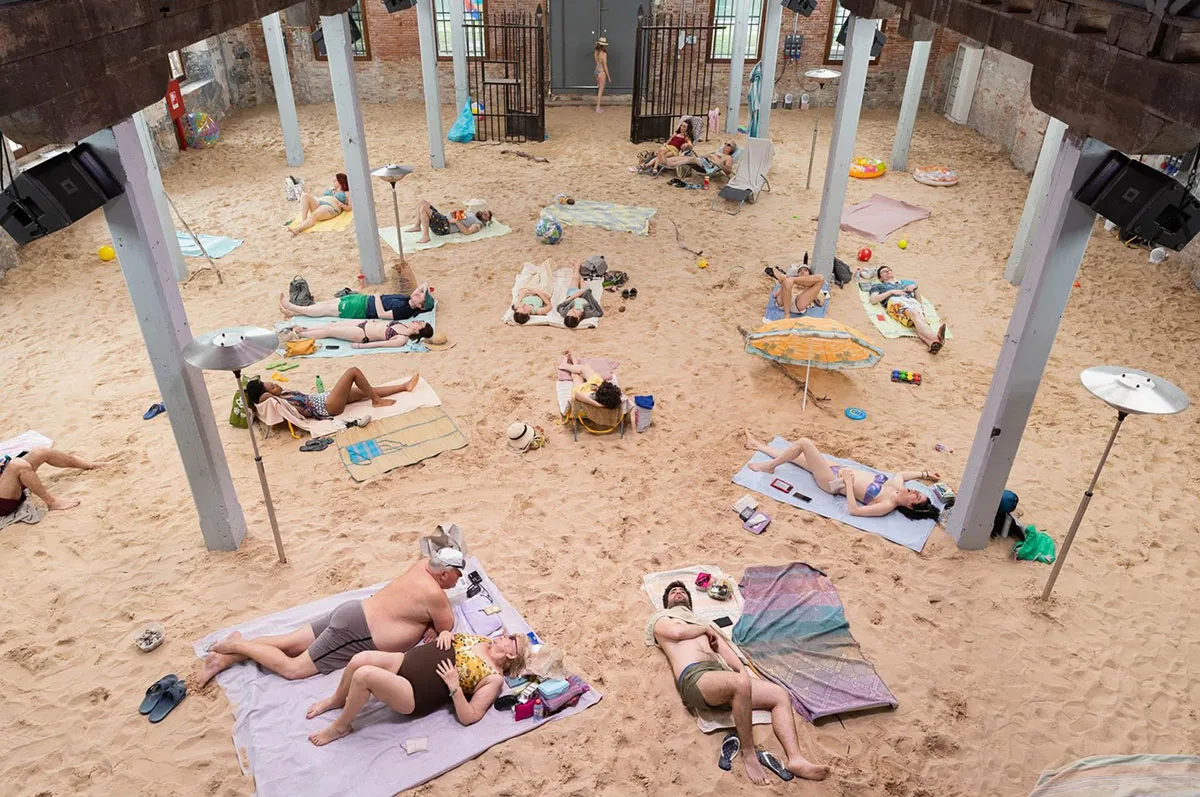 A group of people lying on what appears to be an indoor beach, with speaker boxes hanging from the ceiling