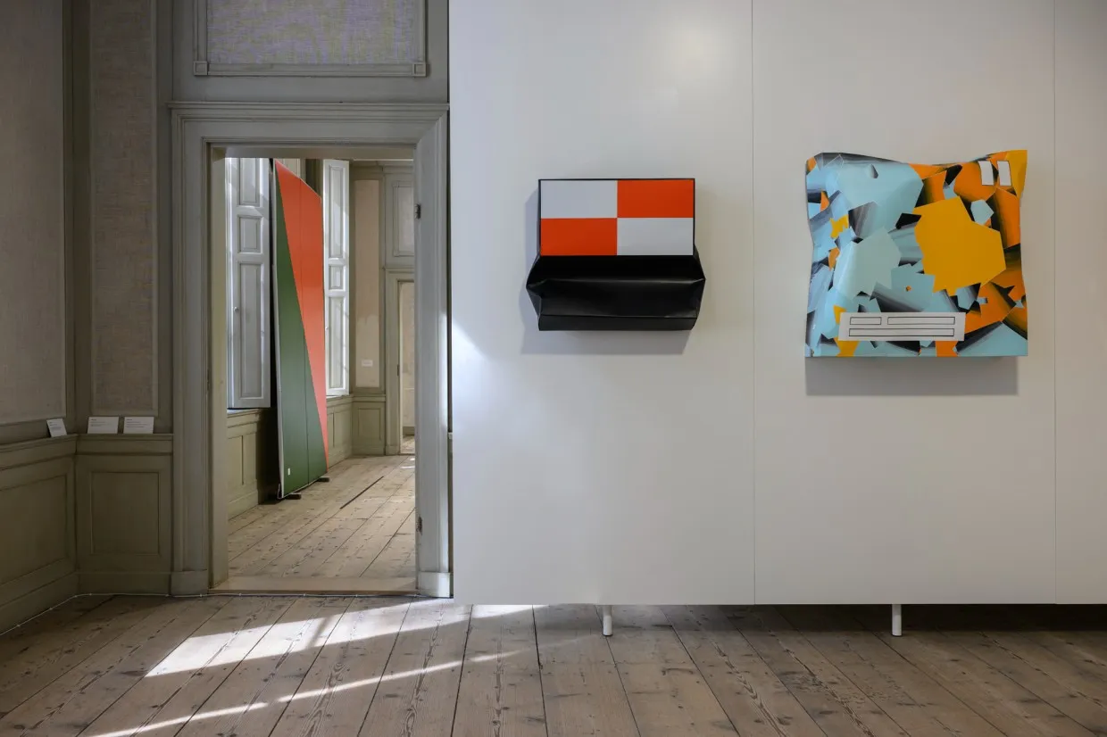 Two smaller square, dented metal sculptures with painted geometric patterns on a wall of the exhibition: one in shades of red, white, and black, and the other in turquoise and orange.