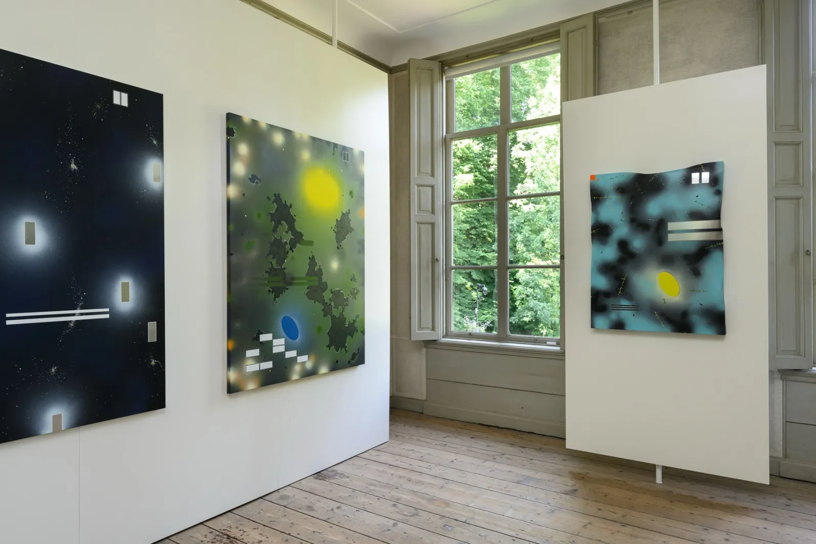Three abstract artworks bathed in natural light beside a window, where trees peek through, within the exhibition space