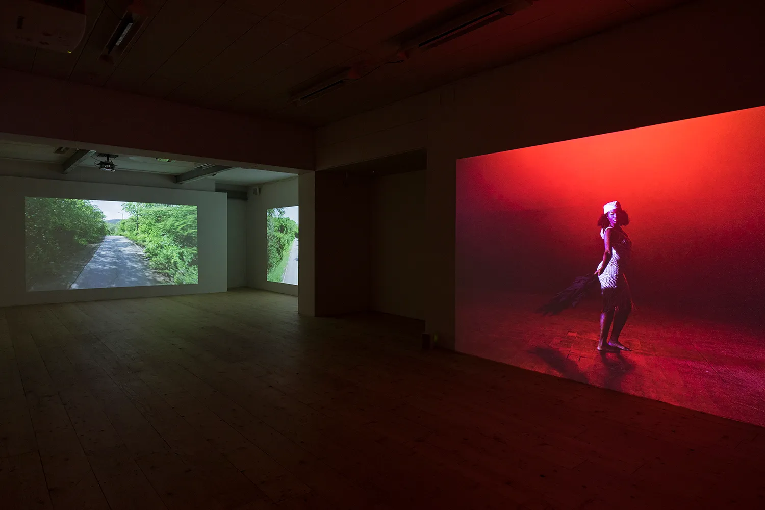 3 projecties op verschillende muren van een lege galerieruimte, twee met vegetatie en een weg; een andere met een in het wit geklede figuur, omgeven door een rode gloed.