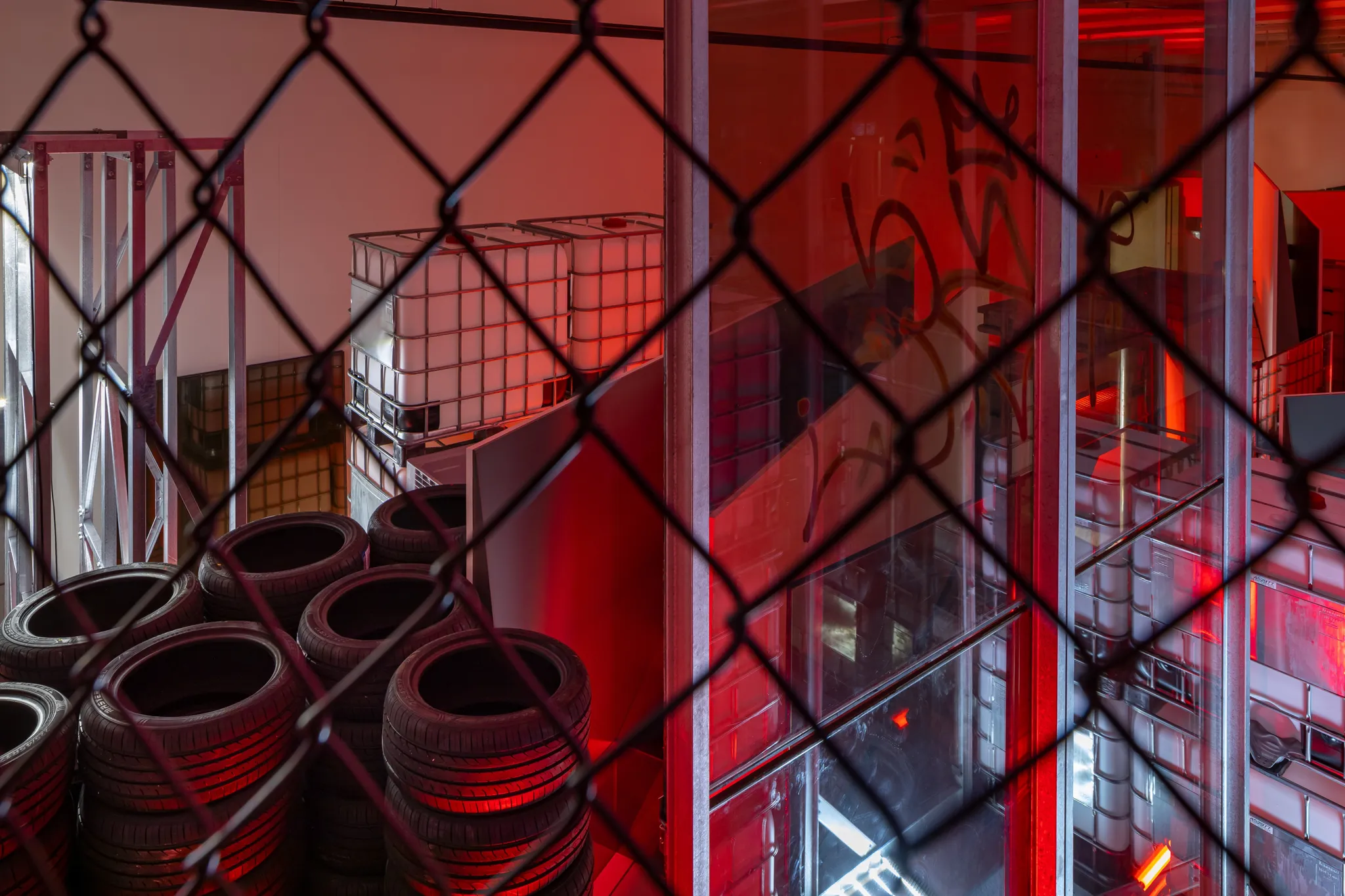 Bovenaanzicht van de tentoonstelling door een metalen hek: autobanden, grote glazen platen met graffiti's, gestapelde watertanks, omgeven door rood licht