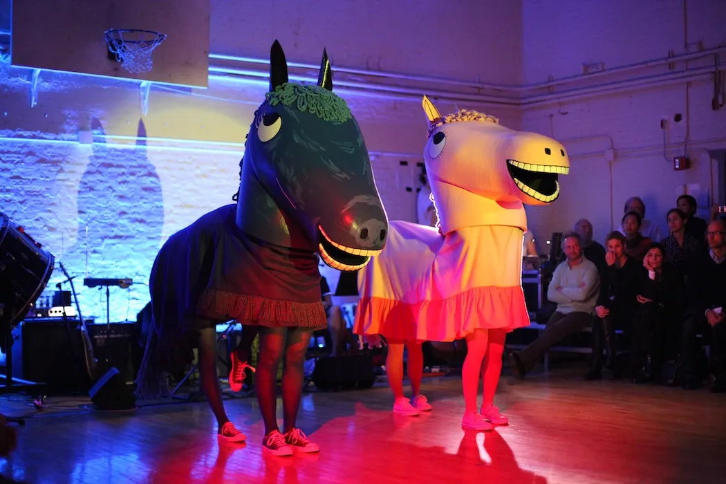 Twee kostuums van lachende cartoonpaarden met performers eronder, aan het optreden voor een publiek op een basketbalveld