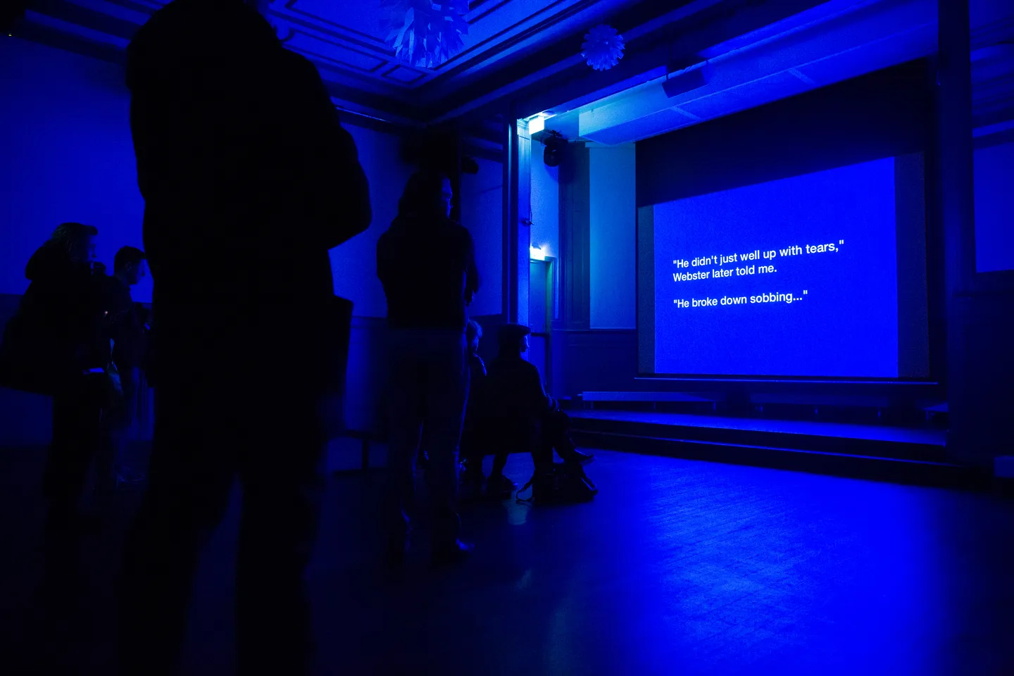 An audience looking at the projection of a text on a bright blue background “He didn’t just well up with tears, Webster later told me. He broke down sobbing…”