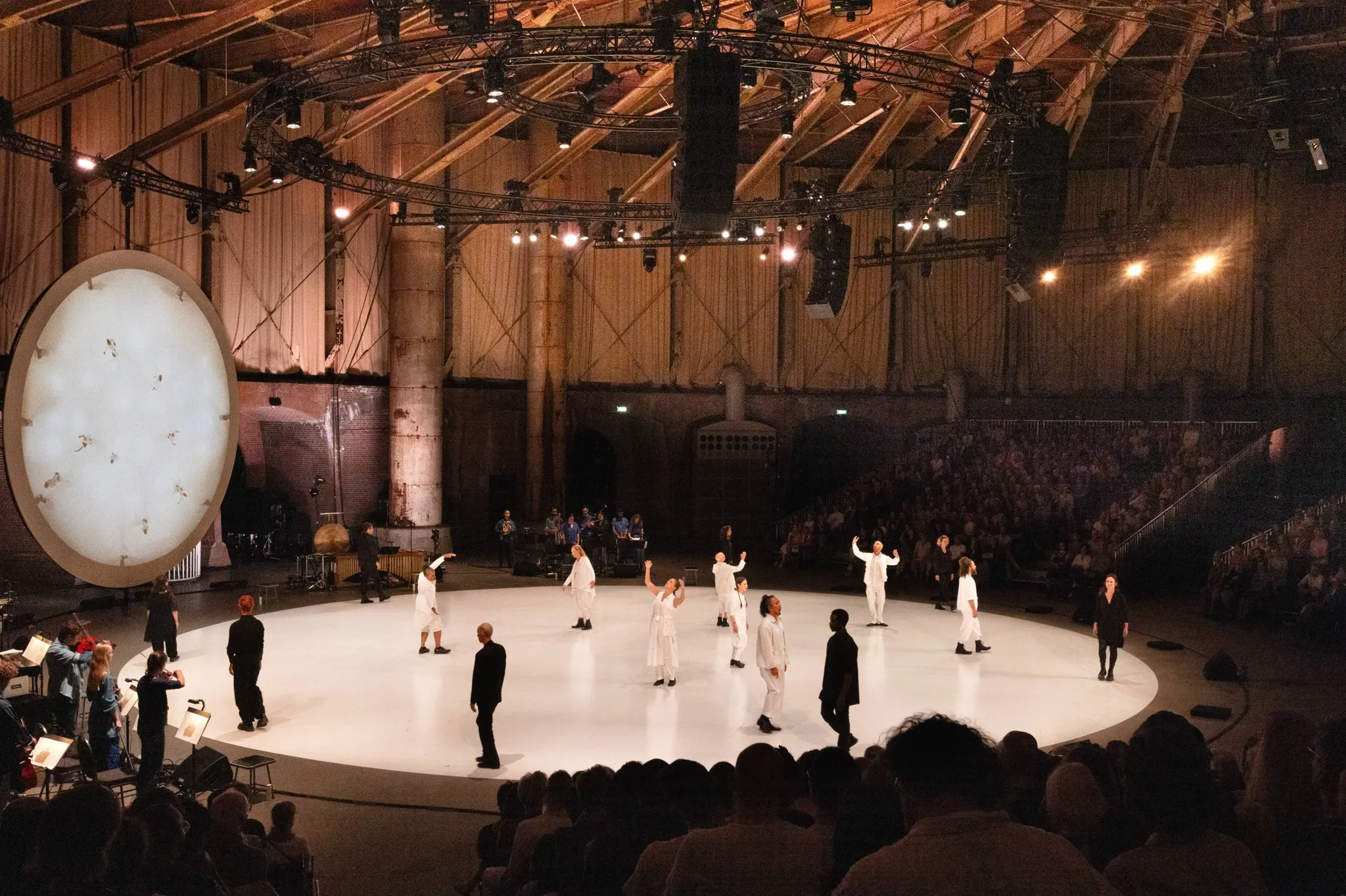 Performers gekleed in wit en andere gekleed in zwart in een cirkelvormige formatie, omringd door muzikanten in een voormalig gashoudergebouw.