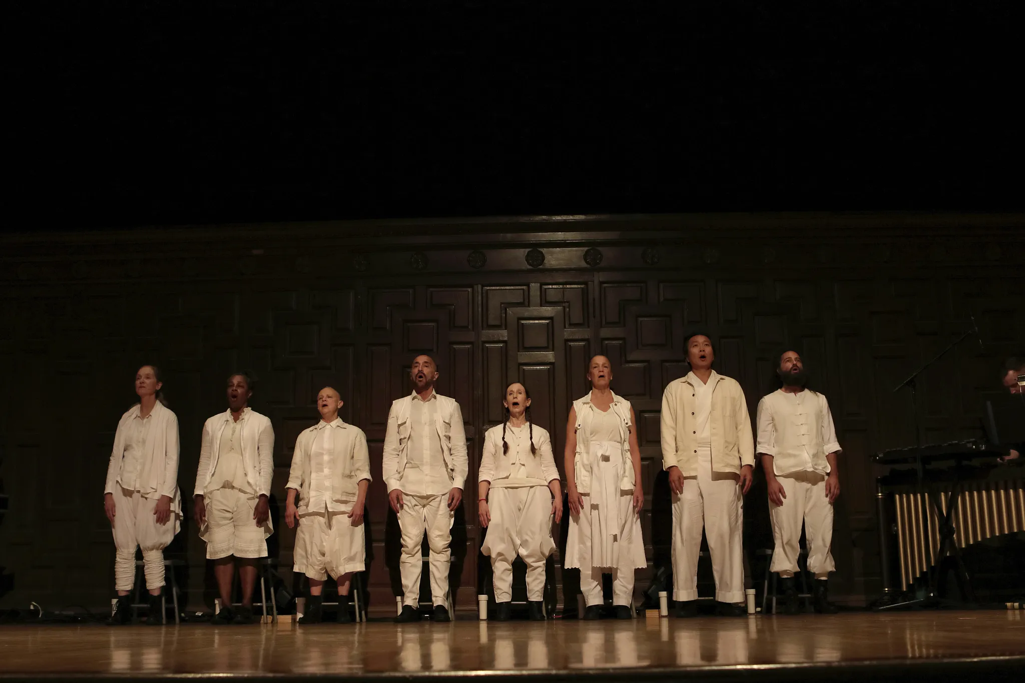 8 personen van verschillende leeftijd, etniciteit, lengte en haardracht zingen in losse witte kleding naast iemand die de marimba bespeelt.
