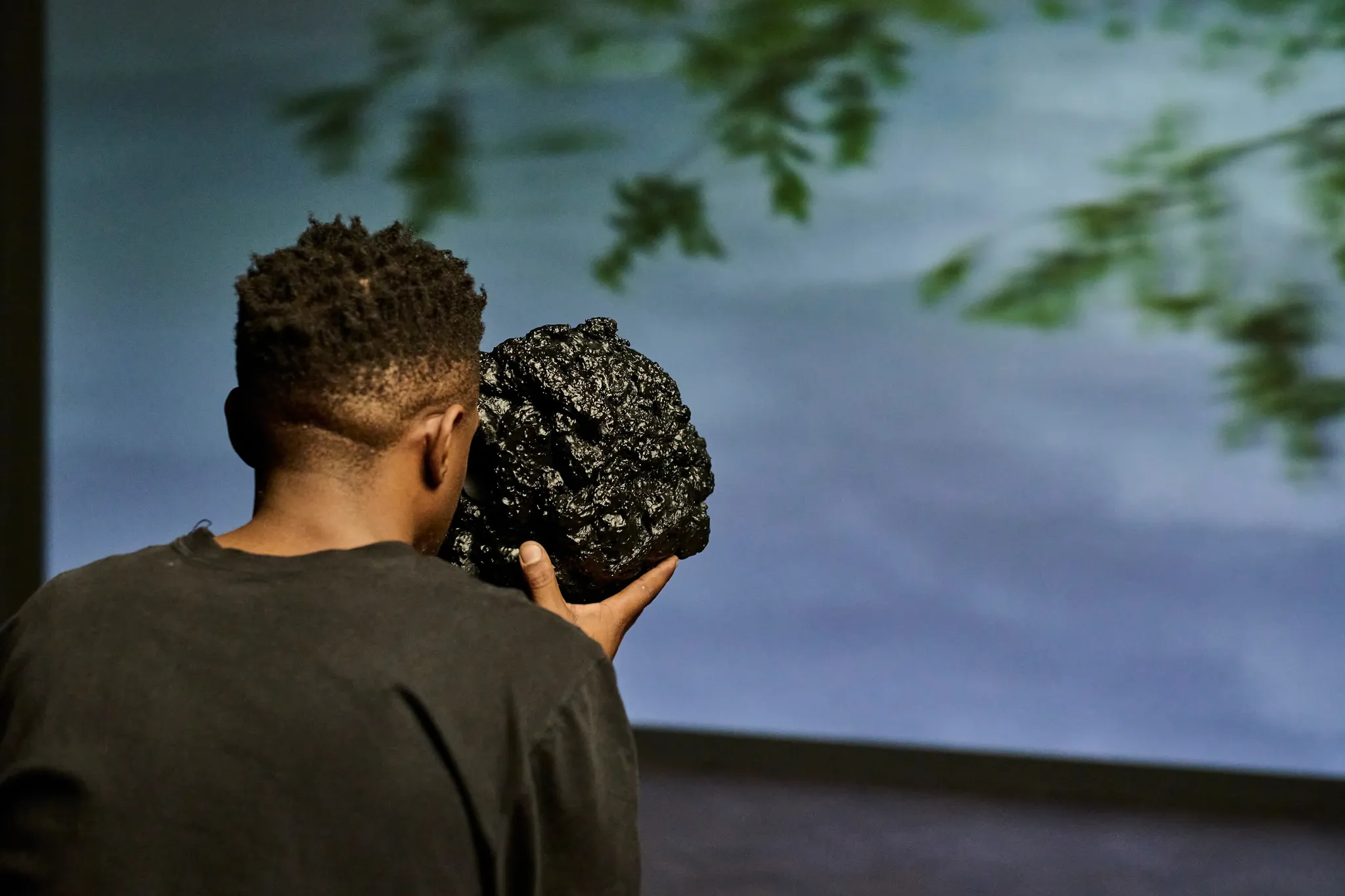 A person with short hair and dark skin looks through a shiny black rock towards a screen projection of blue sky and vegetation