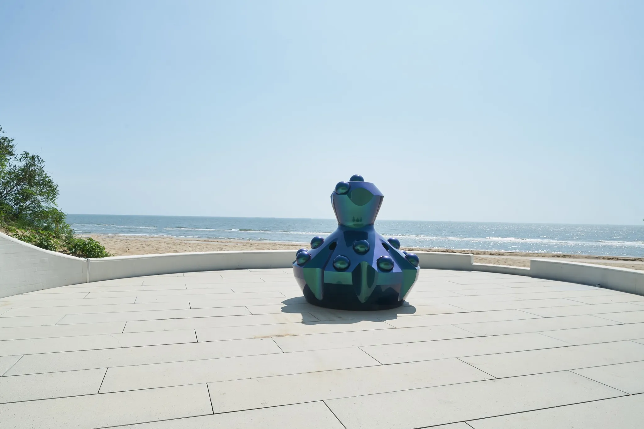 A iridescent-coloured sculpture on a paved terrace facing the beach