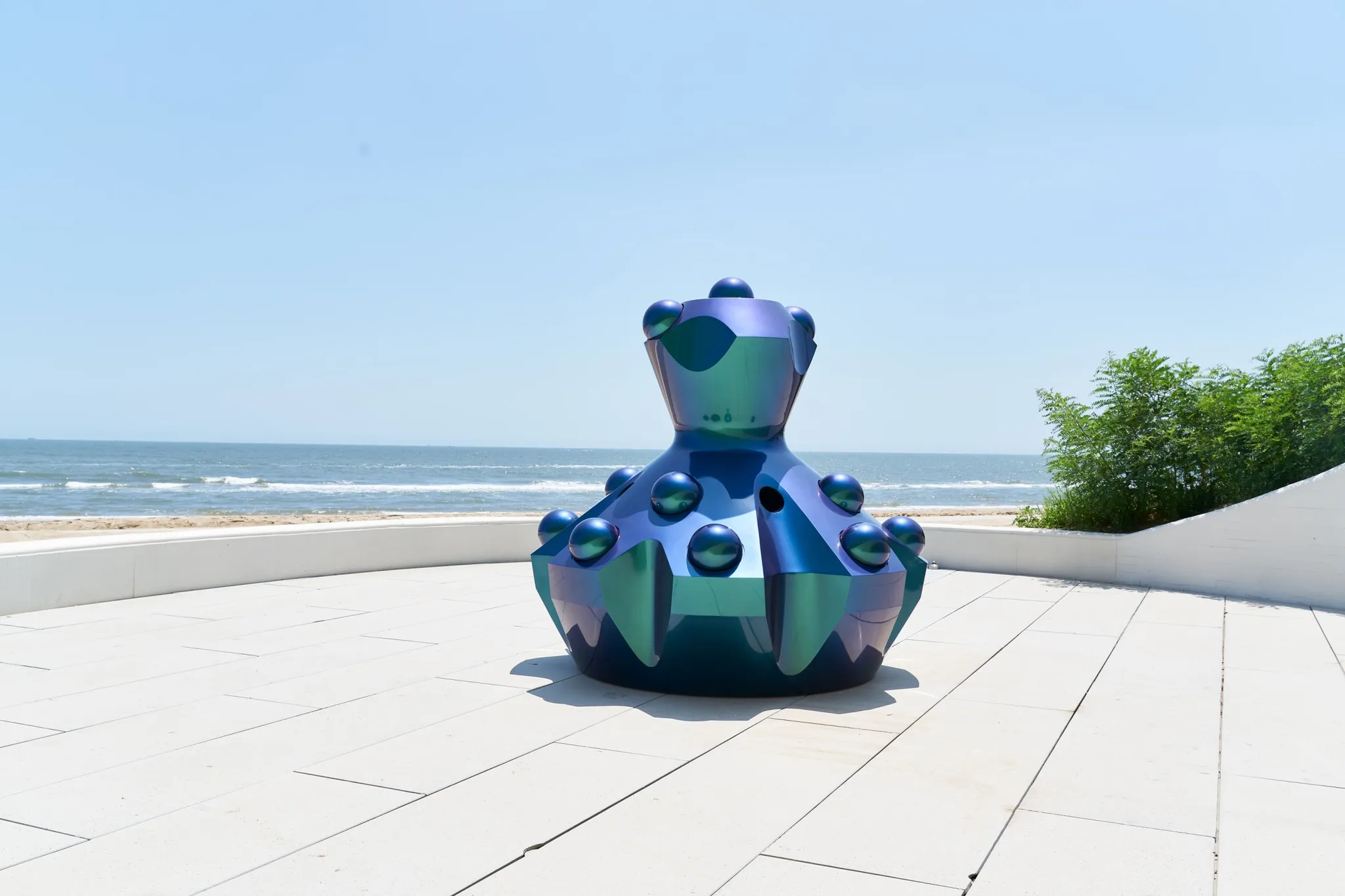 A iridescent-coloured sculpture on a paved terrace facing the beach