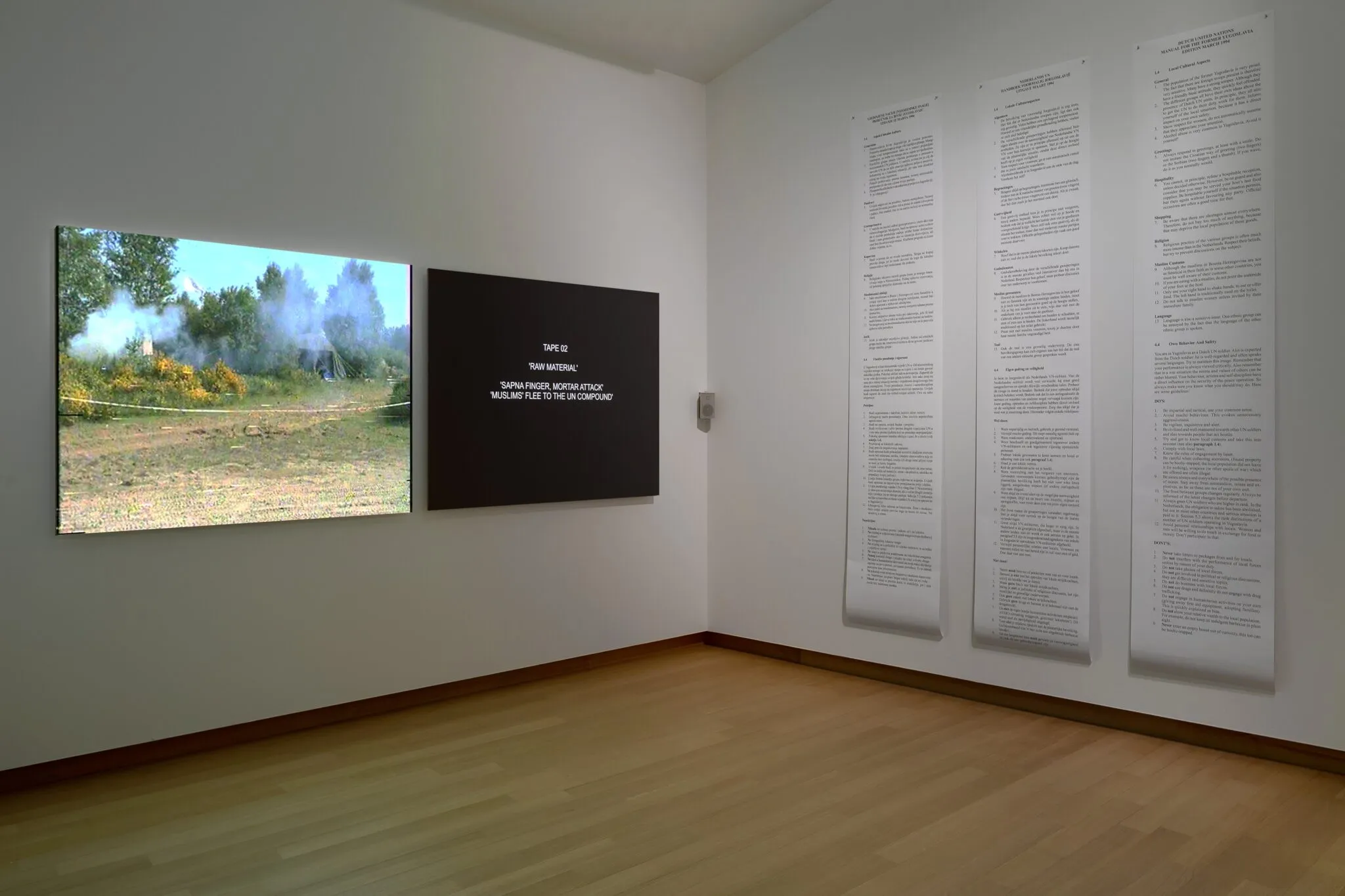 Two screens on a wall and three long sheets of paper with text hung horizontally on the adjacent wall