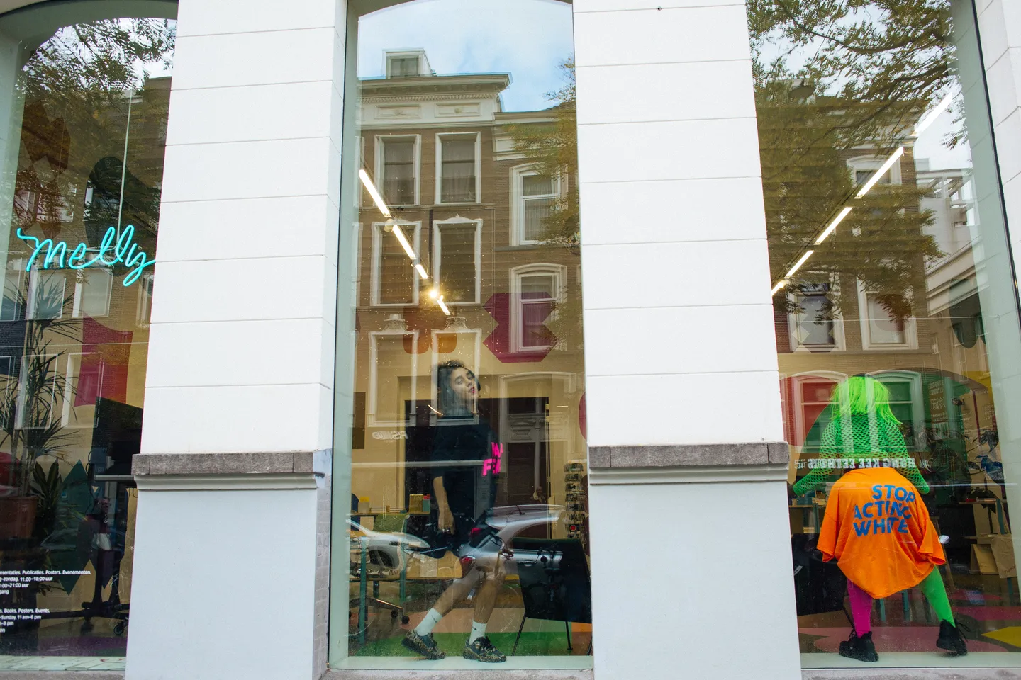 Twee performers dansen op de vensterbanken van Melly. De performer met de groene pruik draagt een oranje t-shirt rond de taille met de tekst 'stop acting white'.