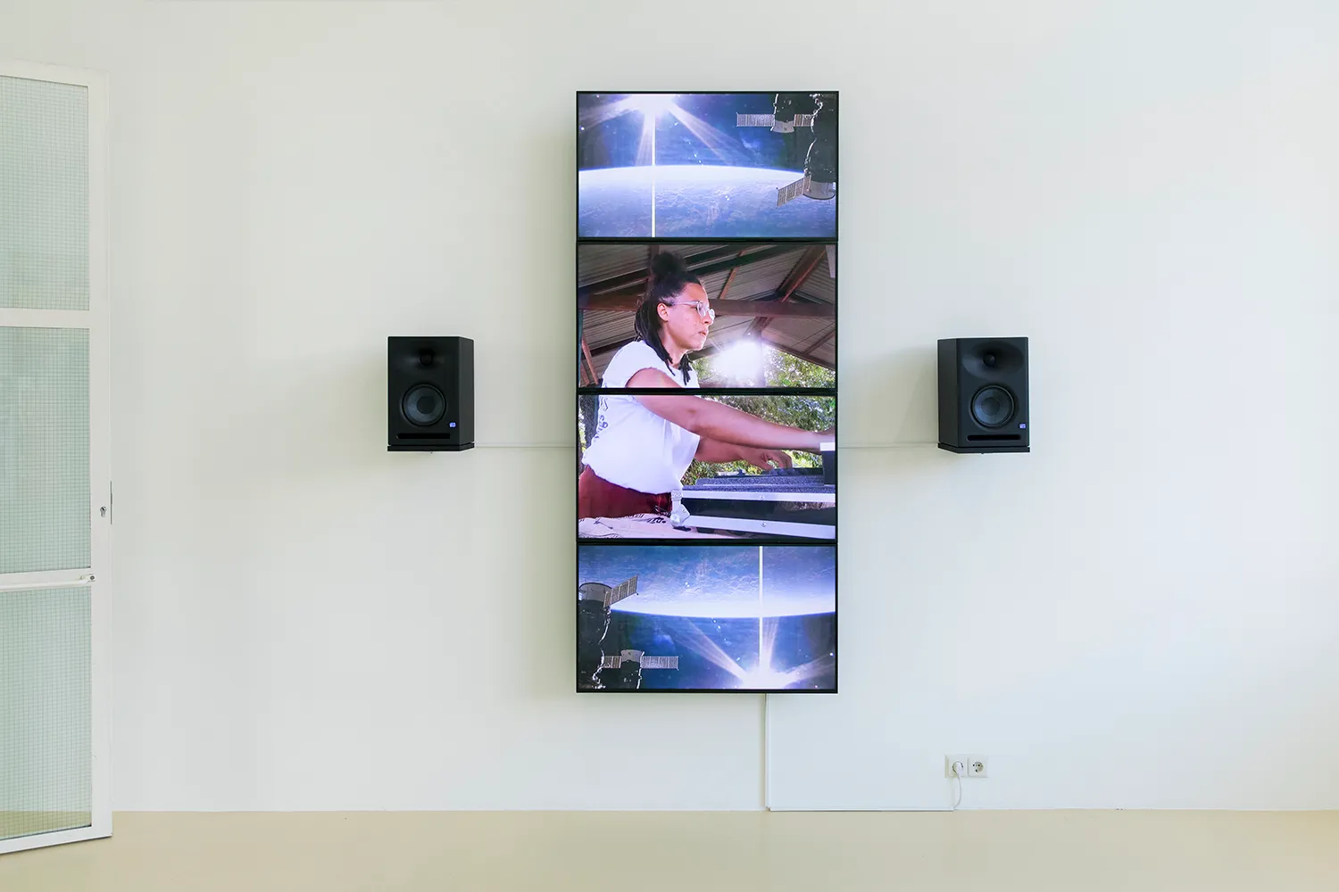 4 flat screens forming a vertical stripe on the wall with 2 speakers. A DJ is mixing in the middle of the screens, surrounded by satellite images of a planet