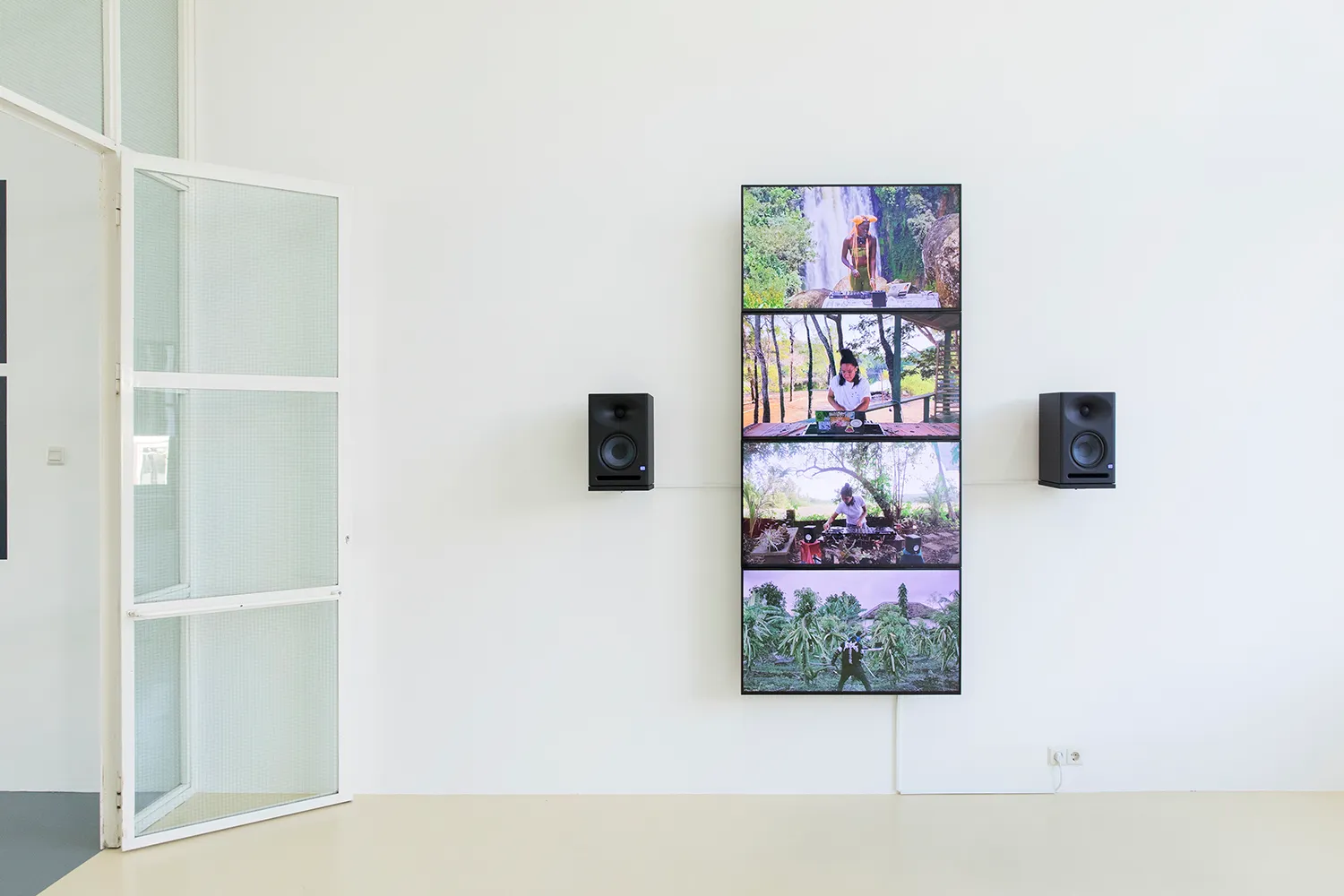 4 flat screens forming a vertical stripe on the wall with 2 speakers. A different DJ is performing on each screen, in front of a tropical background