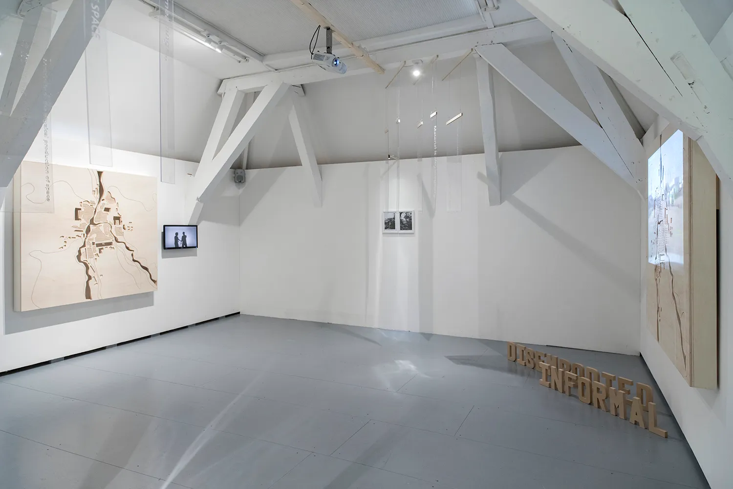 Tentoonstellingsruimte met de 2 gesneden houten planken, klein scherm, 2 fotolijsten, plexiglas stroken die aan het plafond hangen en letters 'disembodied informal' op de vloer