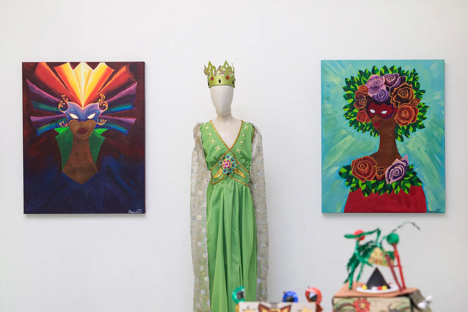 A face-less mannequin with a green dress and a crown stands in the middle of two paintings of a masked person