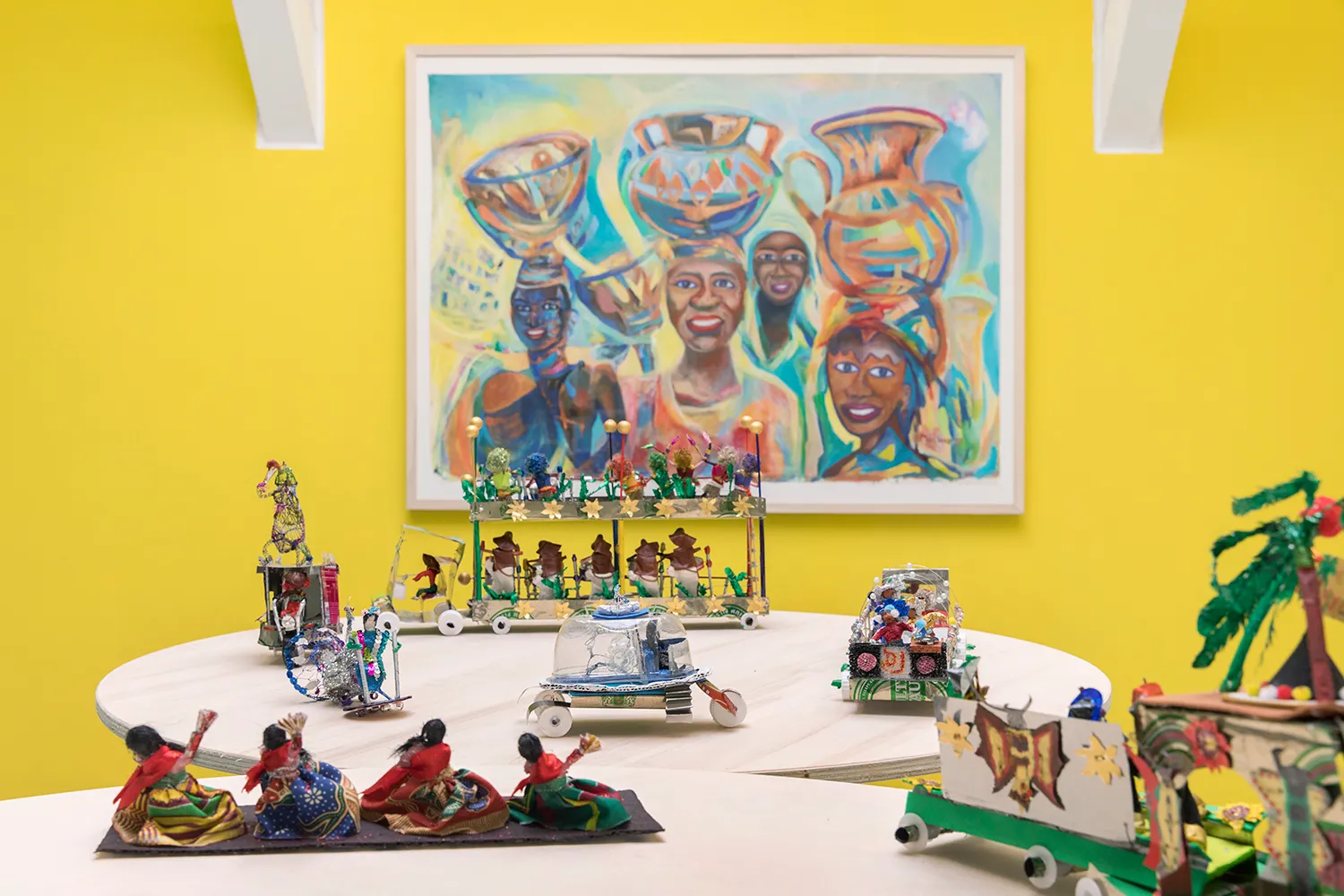 Een tafel met verschillende miniaturen van carnavalsoptochten voor een schilderij van mensen met een donkere huidskleur, lachend terwijl ze containers op hun hoofd dragen.