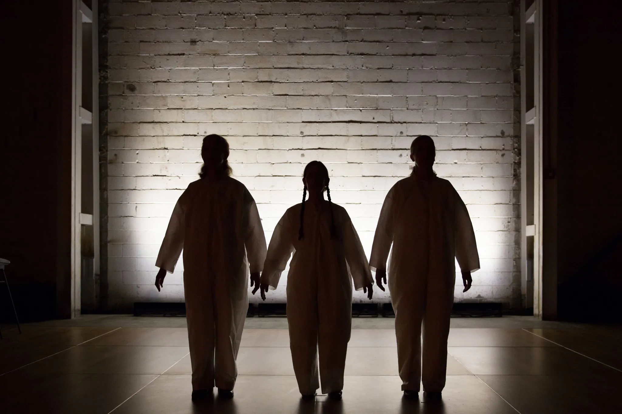 Backlit photograph with 3 silhouettes in white onesies in front of a white bricked wall