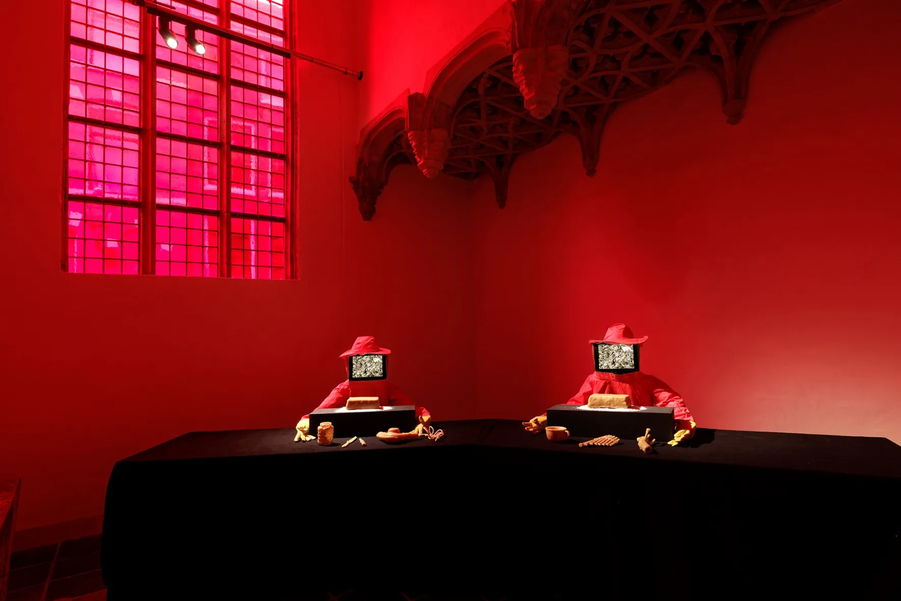 Two red beekeepers outfits with TV screen-covered faces seated at a black table with beeswax objects, illuminated by red light.