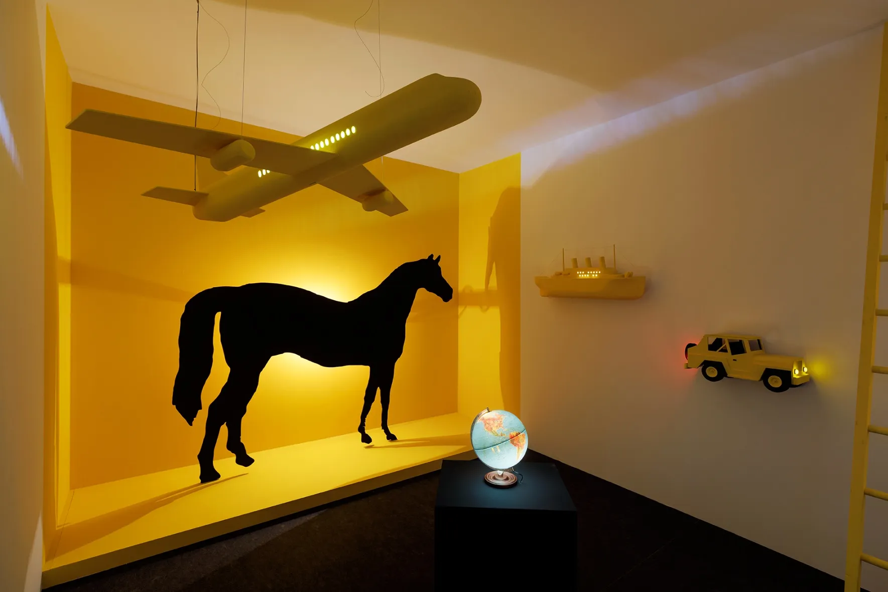 A bright yellow room with props, including a yellow airplane, a black horse silhouette, a yellow boat, and a yellow jeep. A illuminated globe is placed in the center.