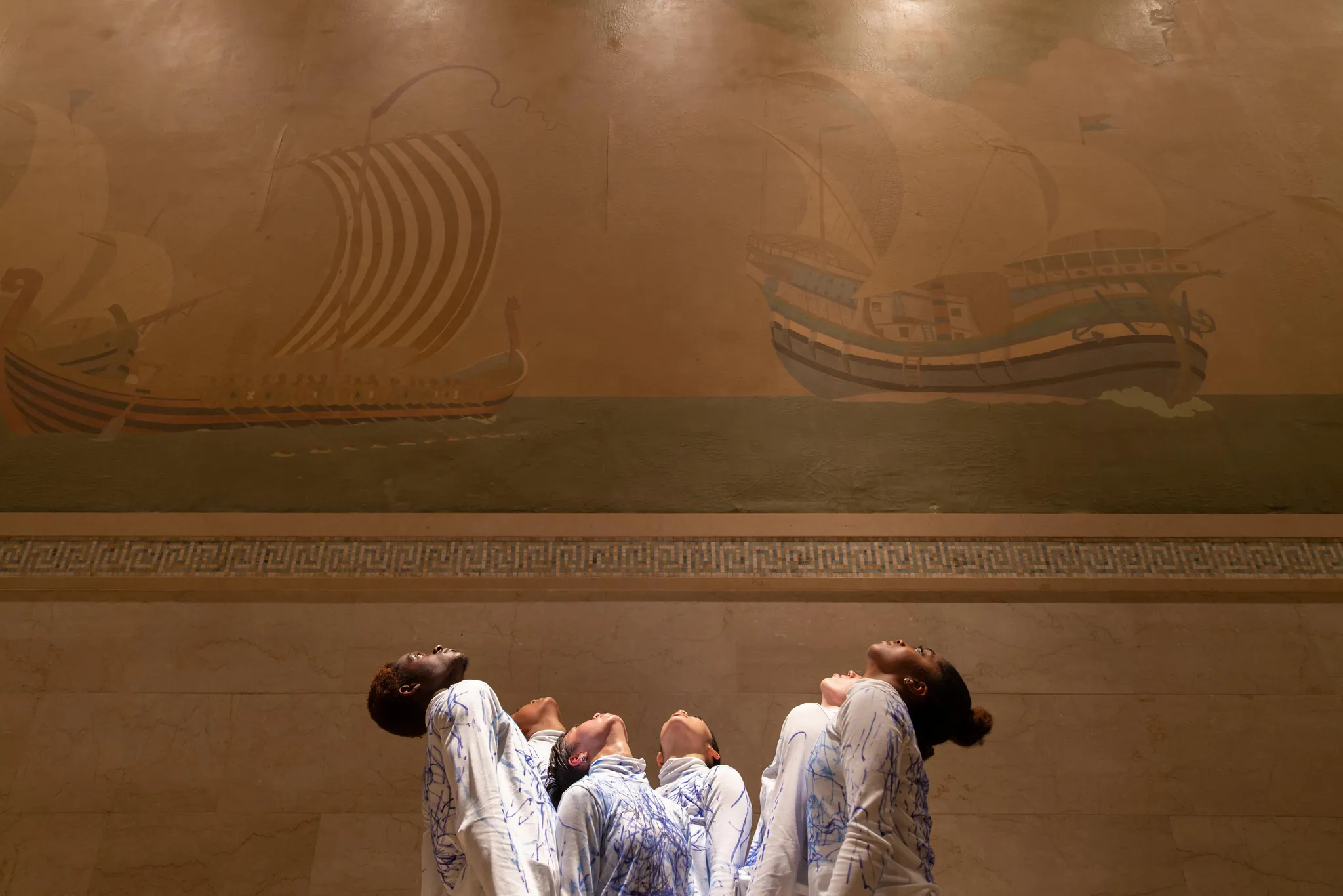 Six performers from diverse ethnic backgrounds, gazing upward in front of a wall painting depicting two ancient ships