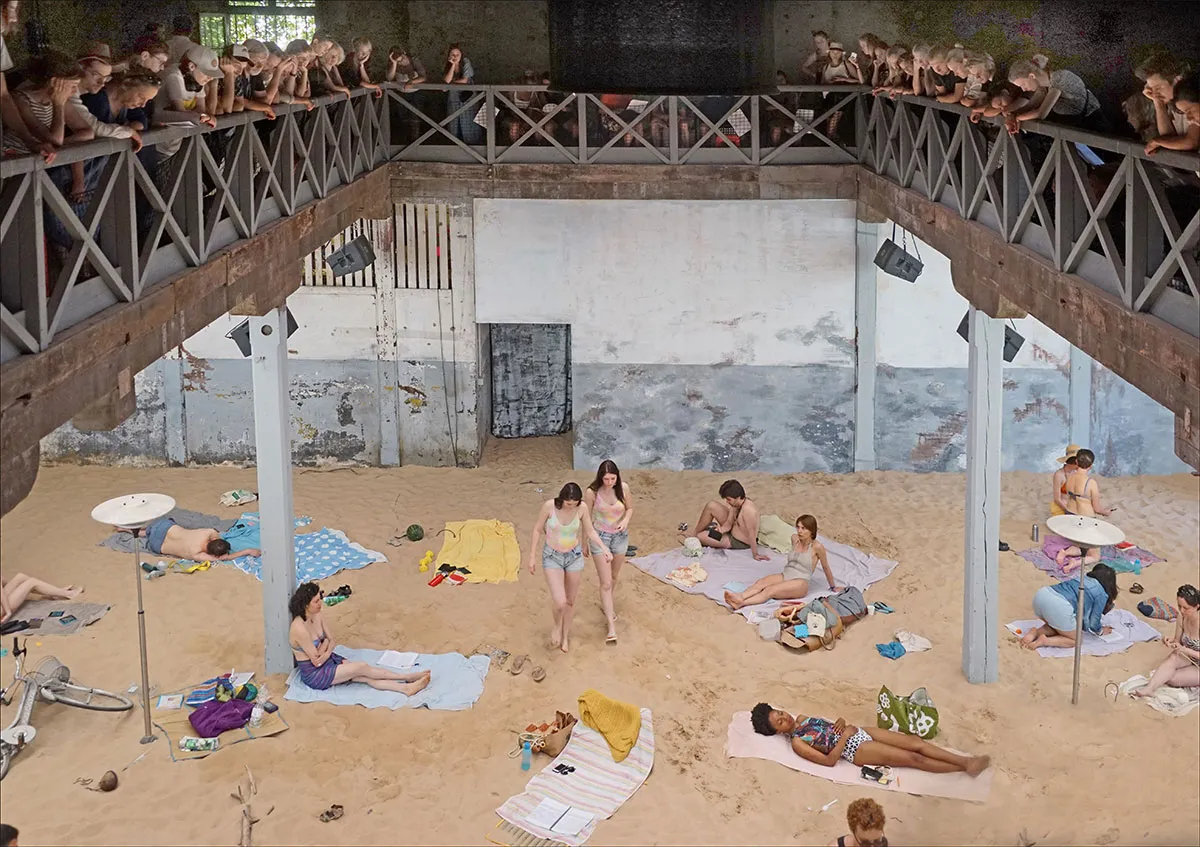 The public looks down from an all-around balcony above performers lying on artificial beach with colourful towels and beach accessories