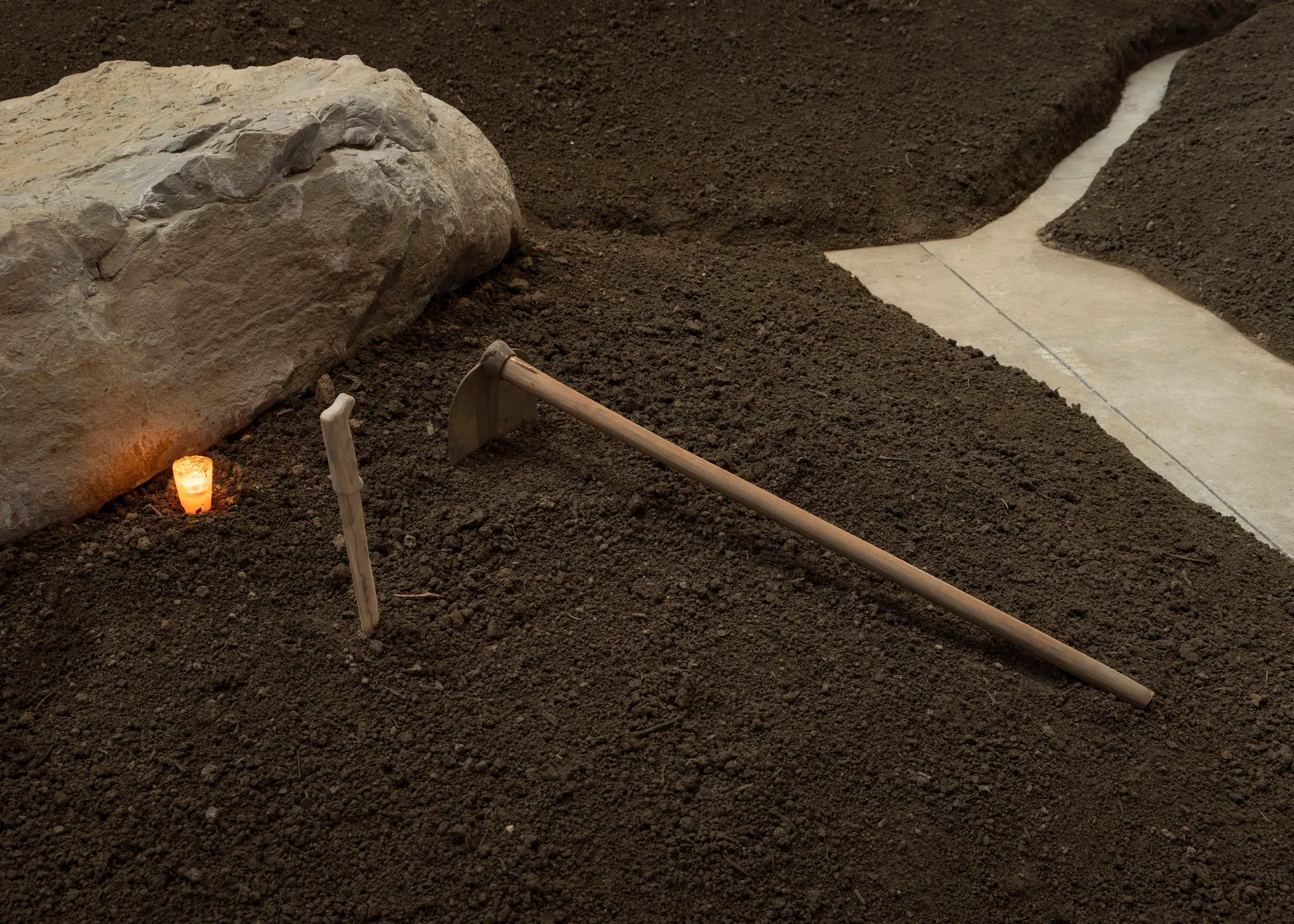 Close up of the installation: a rock, a candle, a wooden knife and a hoe on the brown soil