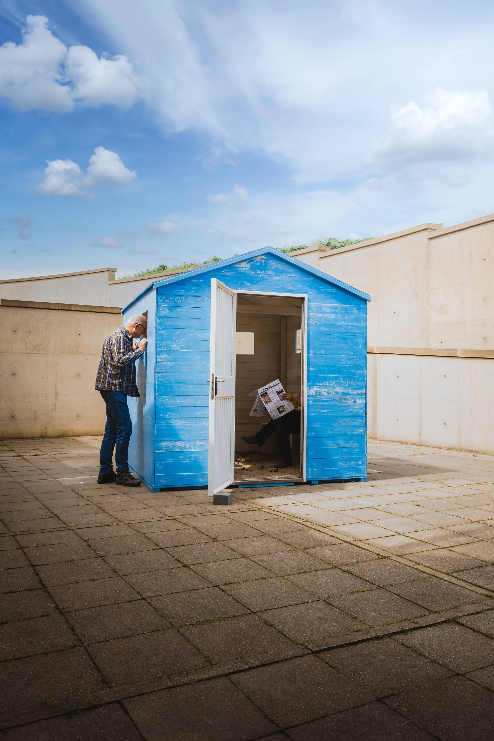 A visitor looks through the window of a blue wooden beach cabin, while another person sits inside, reading a newspaper.